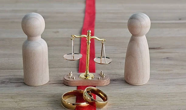 Two wooden figures separated by a red line, with a set of scales and two wedding rings between them